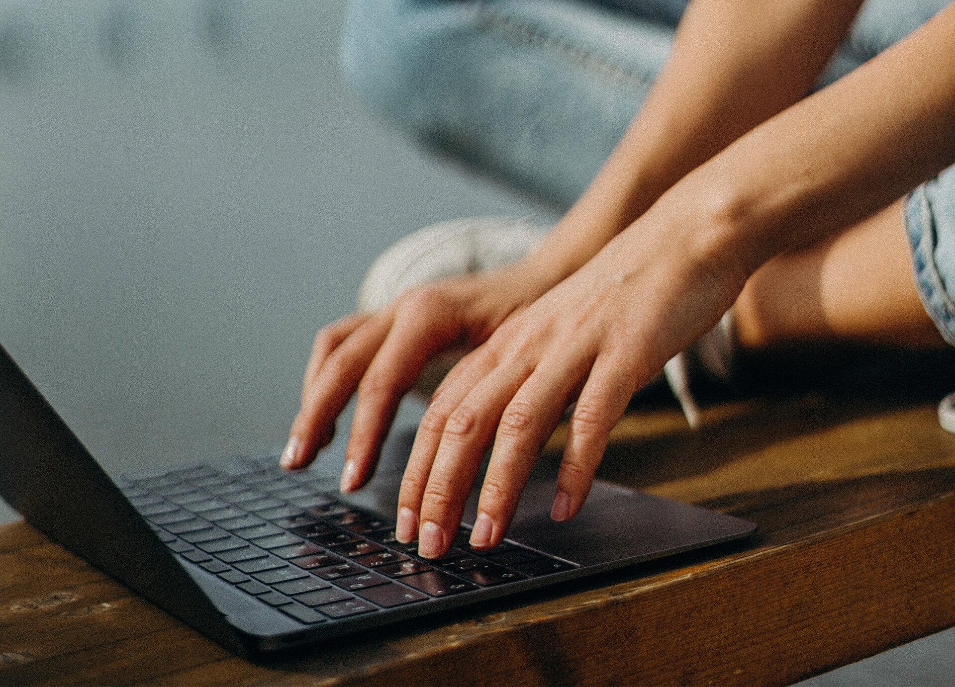 Zdjęcie przedstawia dłonie i nogi osoby siedzącej po turecku na drewnianej ławce i pracującej na laptopie. Pośrodku widoczne są dwie dłonie dotykające klawiatury otwartego laptopa.