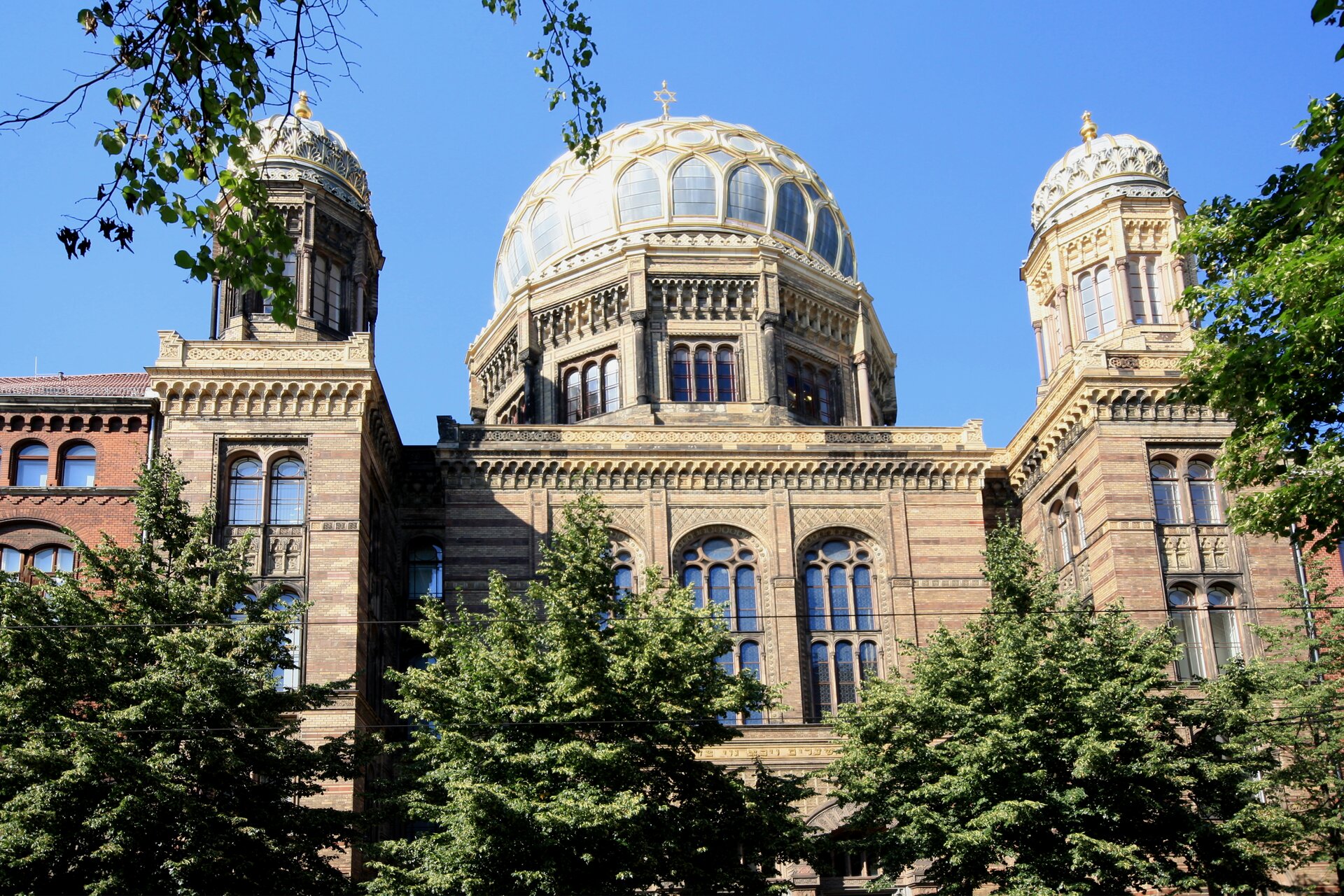 Na zdjęciu murowana synagoga w stylu mauretańskim, wschodnie detale i ornamenty, trzy kopuły, największa z nich znajduję się w centralnej części budynku, dwie mniejsze po bokach. Synagoga umieszczona między innymi budynkami. Na pierwszym planie drzewa.