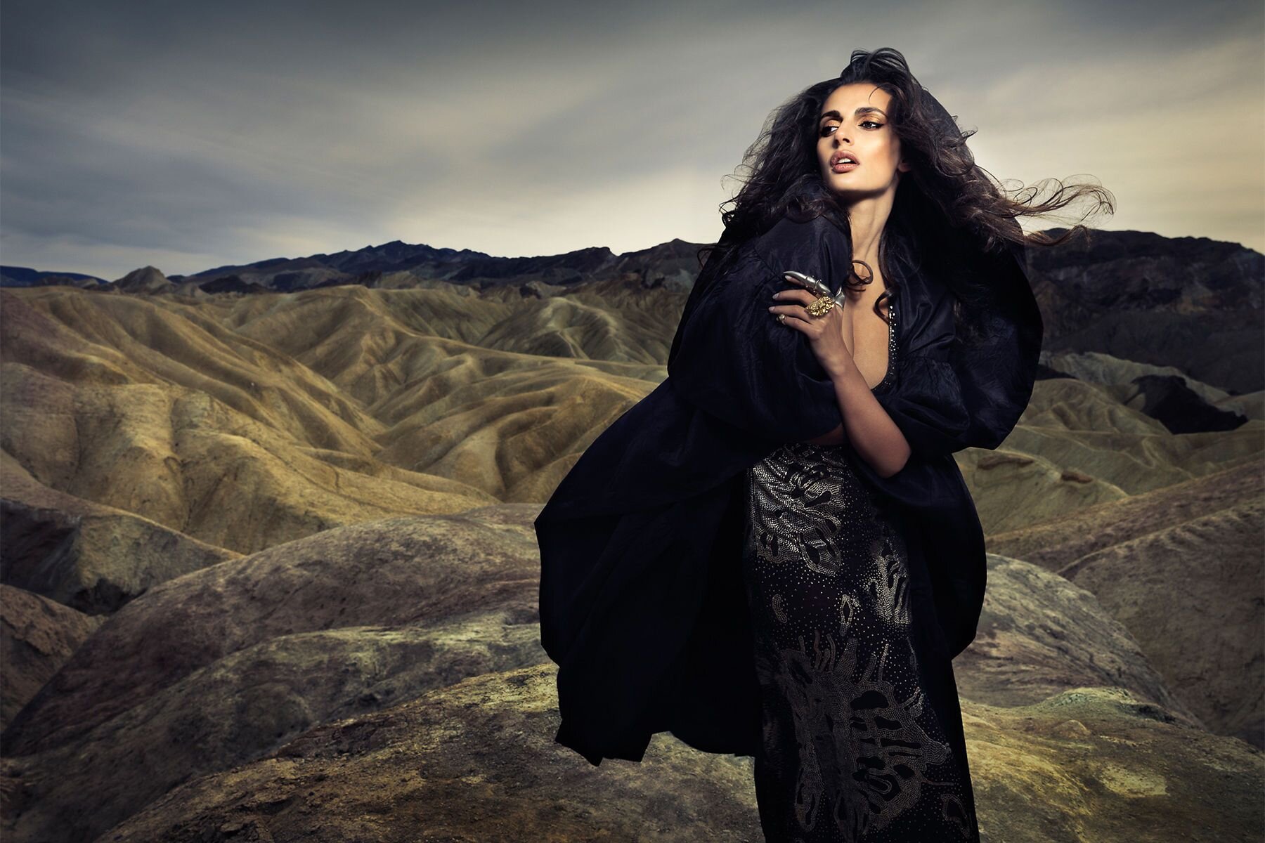 Fotografia przedstawia kobietę na tle naturalnego krajobrazu. Modelka ma na sobie długą, luźną sukienkę w ciemnym kolorze z jaśniejszymi nieregularnymi wzorami. Suknia odsłania jej dekolt, zasłonięty częściowo przez lewą rękę kobiety. Jej ramiona przykryte są obszernym, czarnym materiałem przypominającym pelerynę, sięgającym jej do kolan i rozwianym przez wiatr. Widać spod niego długą szyję kobiety, a także skierowaną w prawo twarz. Jej usta są duże, otwarte – ukazują białe zęby. Makijaż kobiety utrzymany jest w odcieniach brązu – policzki podkreślono ciepłym bronzerem, a oczy czarnymi kreskami na górnej linii rzęs, a także cieniami o neutralnych tonach. Długie, czarne włosy modelki są rozwiane. Na jej lewej dłoni widać biżuterię – srebrną, podłużną  ozdobę pokrywające całą długość palca wskazującego, a także duży, złoty pierścionek. Widoczny za kobietą krajobraz przypomina skaliste wzniesienia w kolorze szarym, żółto‑beżowym oraz czarnym. Nad nimi można zauważyć jasne, zachmurzone niebo.