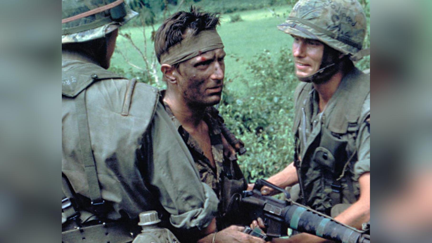 Zdjęcie przedstawia trzech żołnierzy. Dwaj po bokach mają kamizelki i hełmy moro. Żołnierz po środku ma brudną twarz, opaskę na czole, rozpiętą koszulę moro i broń w ręku. W tle jest zielone pole.