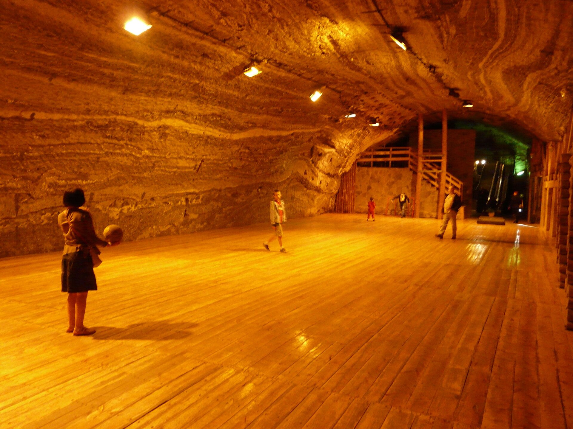 Zdjęcie ukazuje boisko piłkarskie w Komorze Ważyn. To obszerne pomieszczenie o półkolistym stropie. Ściany są nierówne. Mają podłużne bruzdy. Na pierwszym planie stoi kobieta i trzyma piłkę do gry. Nieco dalej stoi chłopiec i mężczyzna. Sala jest oświetlona.