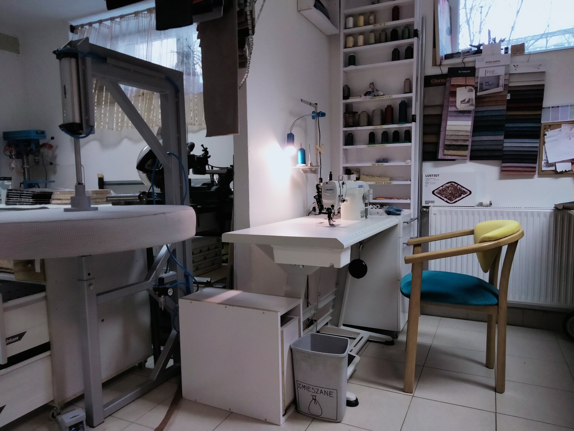 Na ilustracji przedstawiono stanowisko do szycia w pracowni tapicerskiej. Pomieszczenie jest częściowo przedzielone ścianką, przy której znajduje się stanowisko z maszyną do szycia oraz regał ze szpulami nici i próbnikami tkanin. W drugiej części pracowni mieszczą się maszyny tapicerskie.