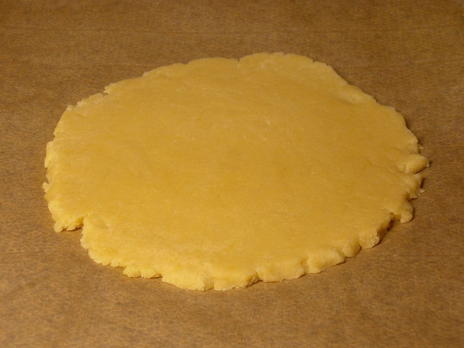 Na fotografii widać rozwałkowane ciasto kruche w formie okrągłego placka leżącego na brązowym papierze do pieczenia. Ciasto jest koloru żółtego. 