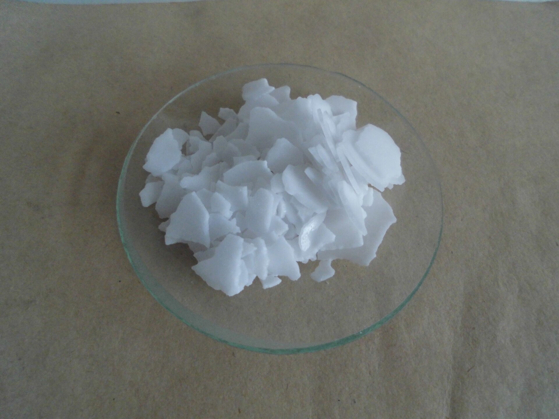 Na zdjęciu na szkiełku zegarkowym znajduje się pewna ilość substancji. Ma postać cienkich, łamliwych, białych płatków.  