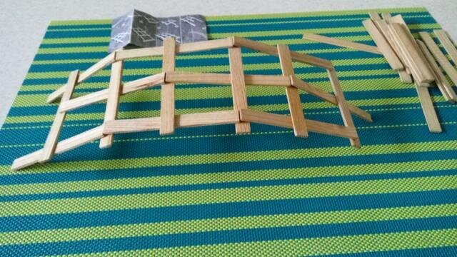 Zdjęcie przedstawia konstrukcję mostu z drewnianych patyczków do lodów. Składa się ona z 6 patyczków, które tworzą długość i 3 tworzących szerokość. Pomysł mostu jest samonośny, ponieważ każdy element (patyczek) łączy się z następnym naprzemiennie; raz nad, raz pod sobą. Dzięki temu powstaje łuk, dla którego nie trzeba już dodatkowych wzmocnień. Model z patyczków leży na stole przykrytym materiałem w paski, ma formę łuku, jest niewielki. Obok niego leżą luźno pojedyncze patyczki do lodów.