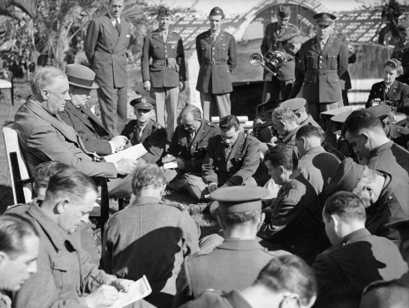 Zdjęcie przedstawia kilkunastu mężczyzn w wojskowych uniformach. Siedzą wokół dwóch mężczyzn siedzących na krzesłach - prezydent USA Franklina D. Roosevelta i premiera Wielkiej Brytanii Winstona Churchilla. Wszyscy piszą coś, albo czytają z kartek. W tle stoi kilku żołnierzy.