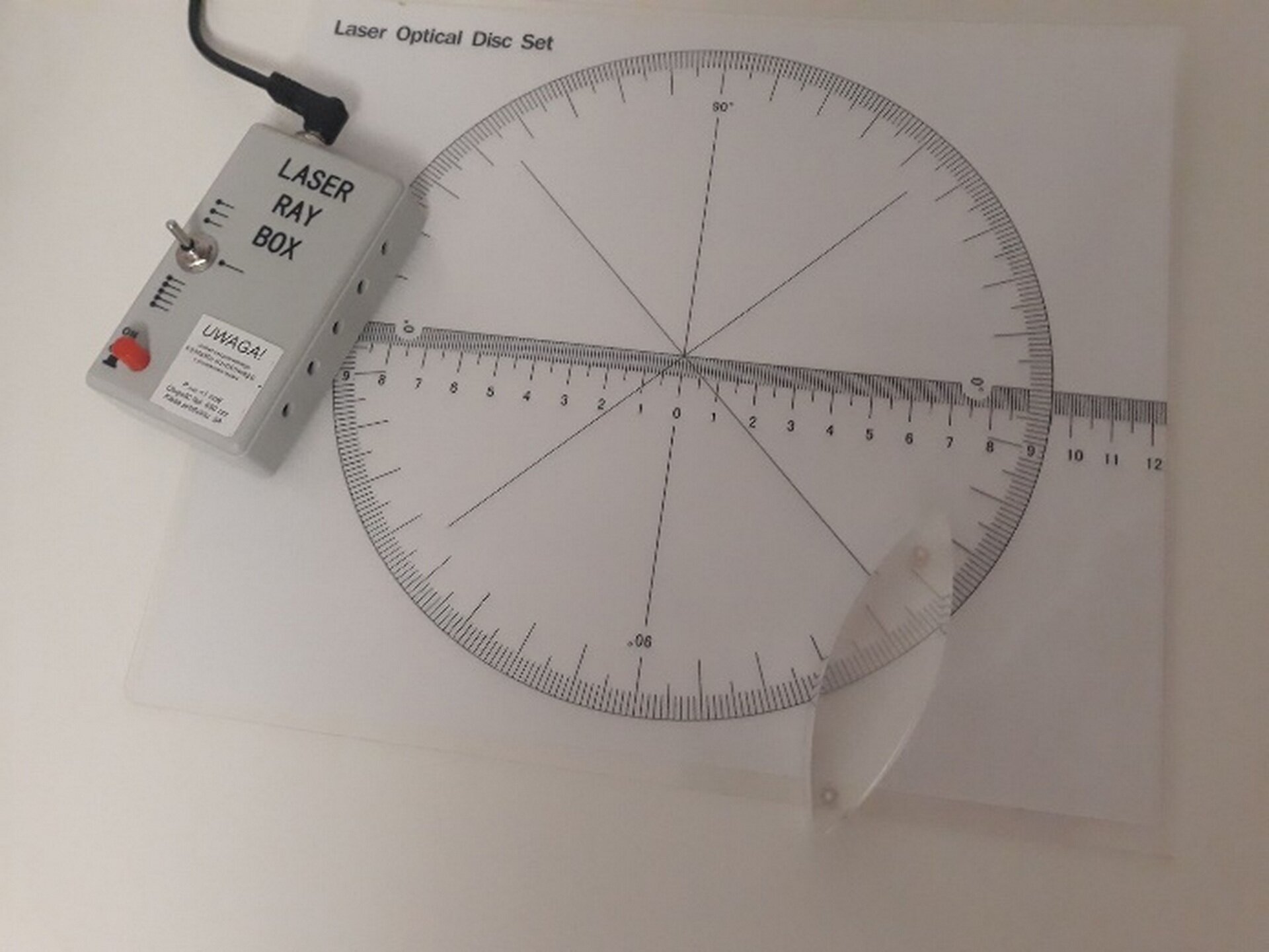 Na ilustracji przedstawiono układ pomiarowy wykorzystywany do określania zdolności skupiającej soczewki. Jest to wydrukowana na papierze okrągła skala z podziałką w stopniach oraz skalą liniową widoczną wzdłuż średnicy okręgu. Ponadto w lewym górnym rogu zdjęcia zaprezentowano oświetlacz emitujący jedną, trzy lub pięć wiązek lasera. W prawym dolnym rogu natomiast, umieszczony jest przezroczysty kształt, wypukły po obydwu stronach, czyli soczewka dwuwypukła.  