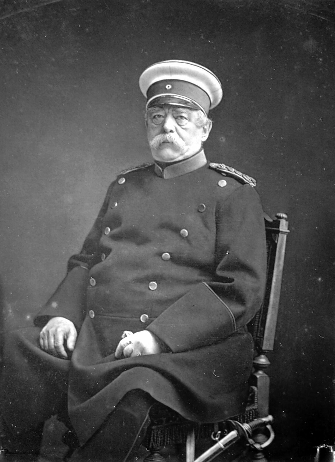 Fotografia czarno-biała przedstawiająca portret Otto von Bismarck'a. Postać siedzi na krześle ubrana w mundur, na głowie ma kapelusz. Spod nakrycia głowy można dostrzec siwe włosy.