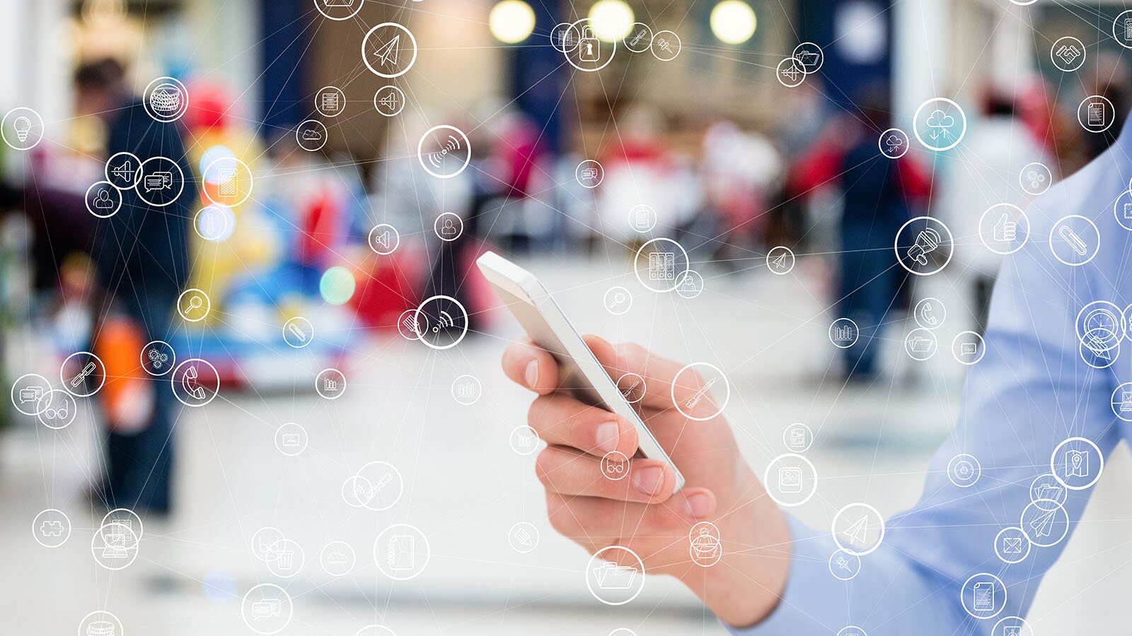 Grafika przedstawia osobę trzymającą w dłoni telefon, wokół którego widoczne są połączenia z różnymi sieciami i urządzeniami. W tle rozmazany obraz ludzi na szerokiej ulicy lub placu.