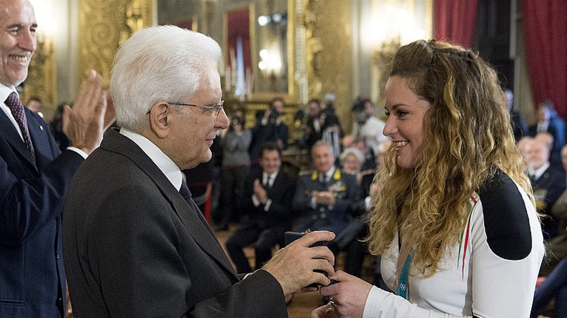 Zdjęcie przedstawia włoską mistrzynię olimpijską w snowboardzie Michelę Moioli. Odbiera nagrodę z rąk siwego, starszego mężczyzny w okularach. W tle widoczni są ludzie, którzy biją brawo.