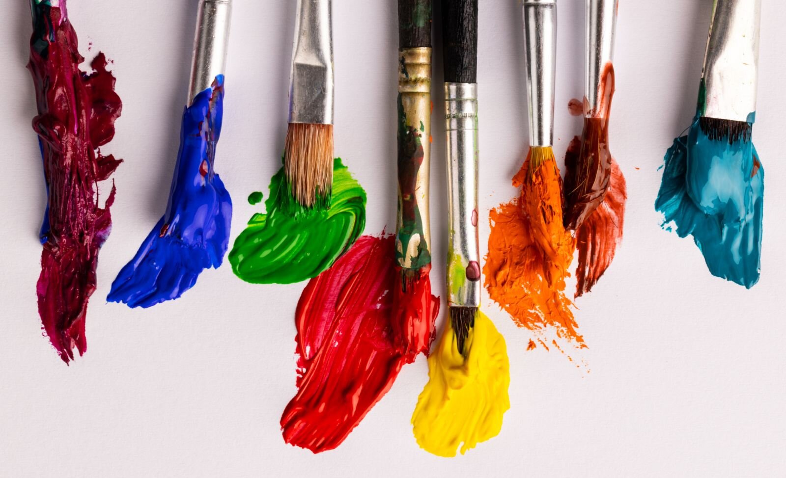  Na zdjęciu kilka grubych pędzli umoczonych w gęstej farbie leży na stole. Farba dotyka blatu stołu pozostawiając plamy farb w różnych kolorach; ciepłych i zimnych. Pędzle są różnej wielkości i grubości, każdy jest zanurzony w farbie, niektóre aż po metalową skuwkę.