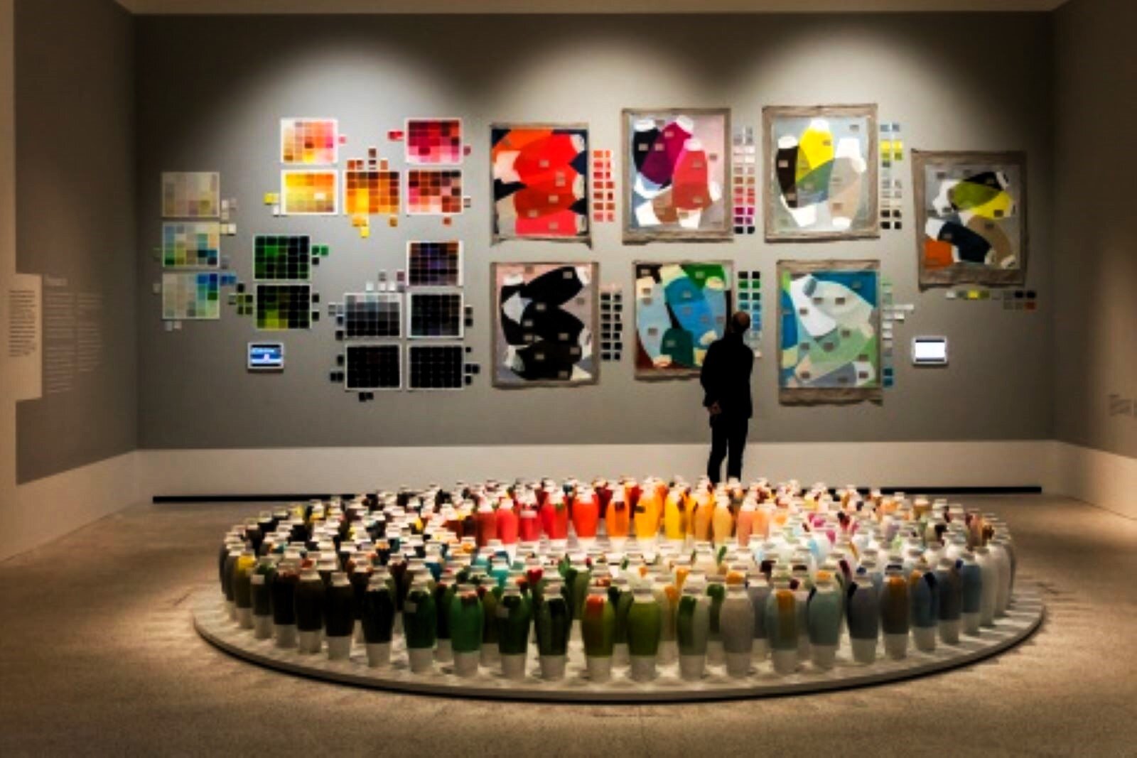 Fotografia przedstawia kompozycję abstrakcyjnych obrazów na ścianie galerii oraz instalację z jednakowych form w kształcie okręgu, ustawioną obok na podłodze. Elementy zarówno jednej, jak i drugiej aranżacji tworzą gamy barwne, ciepłe, zimne, gorące - współistnieją w jednej przestrzeni tworząc uzupełniające się kompozycje na temat koloru.