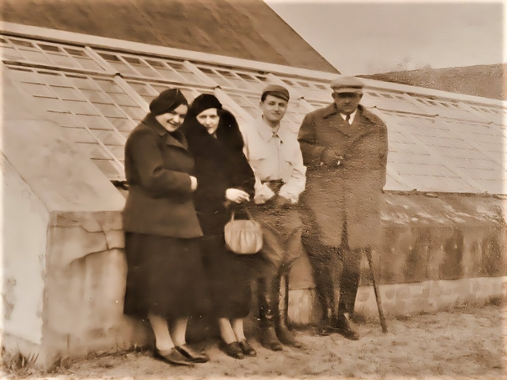 Zdjęcie przedstawia cztery osoby stojące na tle niskiej szklarni. Pierwsza osoba od lewej, kobieta, ubrana jest w długą spódnicę za kolana, skórzane niskie buty, grubą kurtkę i beret. Kobieta uśmiecha się. Obok niej stoi druga kobieta, ubrana w niskie, skórzane buty, płaszcz i beret. W ręku trzyma małą, jasną torebkę. Obok niej stoi młody mężczyzna w wysokich, sięgających do kostek, butach skórzanych, luźnych spodniach, białej koszuli i czapce. Obok niego stoi czwarta postać, starszy mężczyzna. Ma on wysokie, skórzane buty, luźne spodnie wpuszczone w buty, długi płaszcz. Na głowie ma czapkę, prawą dłoń trzyma na guziku płaszcza, a lewą podpiera na lasce. Zdjęcie jest sepią.