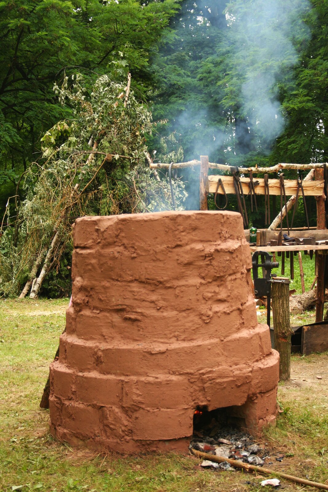 Zdjęcie przedstawia zrekonstruowaną dymarkę, czyli dawny piec hutniczy z cegieł i gliny, w którym wytapiane było żelazo z ich rud. Kształtem przypomina ona duży komin zwężający się ku górze. W dolnej części dymarki widoczny jest prostokątny otwór, w którym umieszczano węgiel drzewny i przez który przedostawało się powietrze niezbędne do podtrzymania ognia. Z górnej części dymarki wydobywa się dym. W tle zdjęcia znajdują się zielone drzewa.