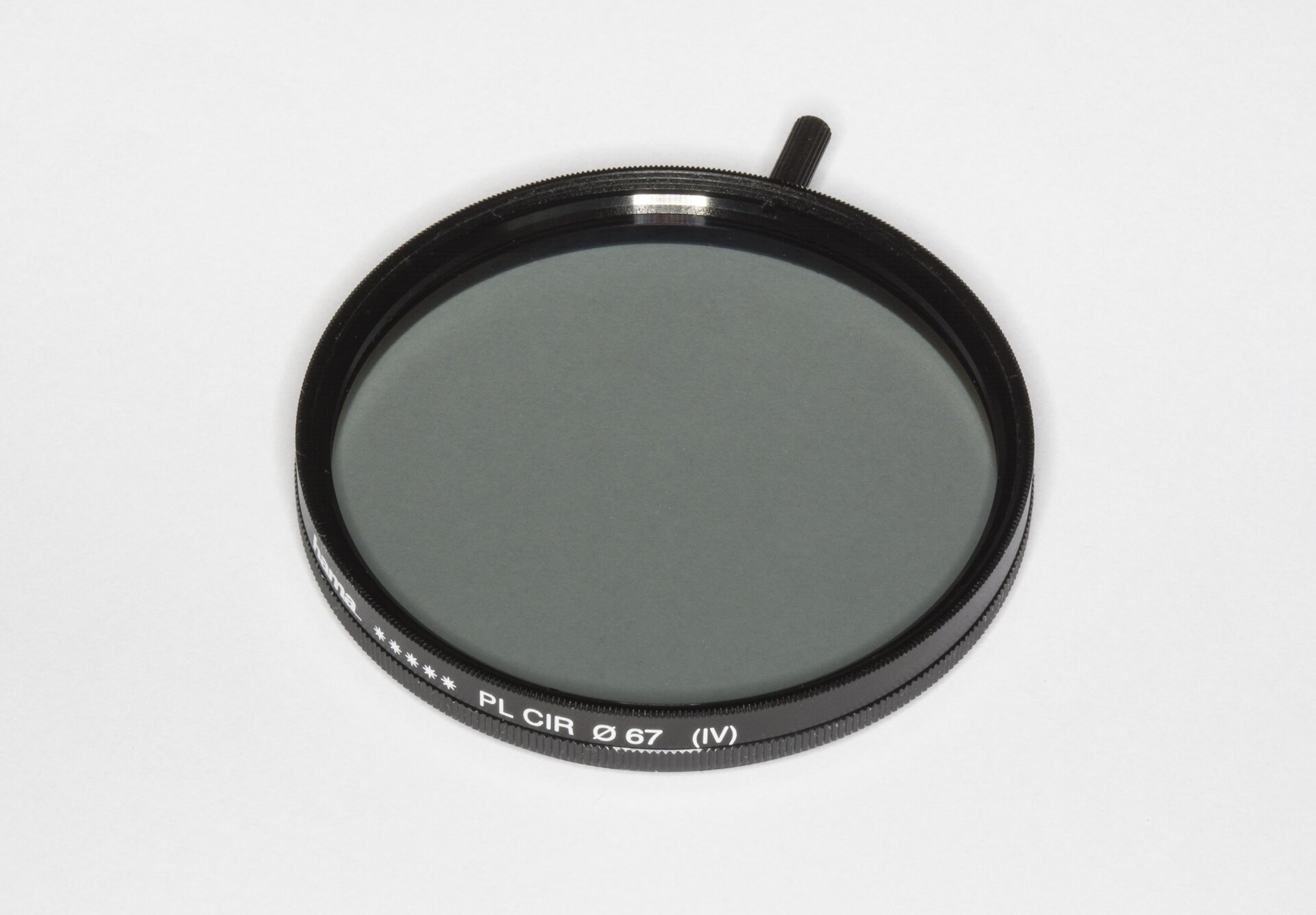 Zdjęcie przedstawia przykładowy filtr polaryzacyjny. Widoczny jest okrągły przedmiot z czarną obwódką i szarym szkłem w środku, U góry widoczny jest krótki cylindryczny uchwyt.