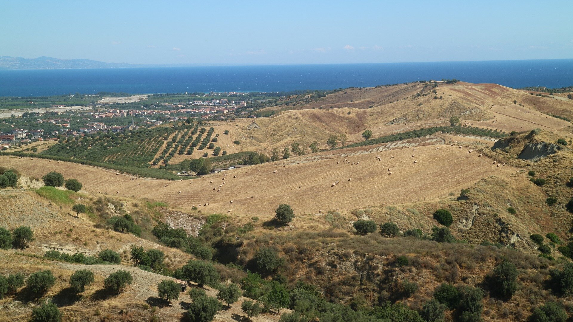 Fotografia przedstawiająca widok ze wzgórza na rozległy, pofalowany teren rolniczy. Na stokach pagórków rozciągają się pola uprawne, na których znajdują się bele sprasowanej słomy. Dookoła nich drzewa oliwne rosnące równo w rzędach. Na drugim planie, nad morzem, położona niewielka osada. W tle morze, a w oddali pasmo wzniesień.