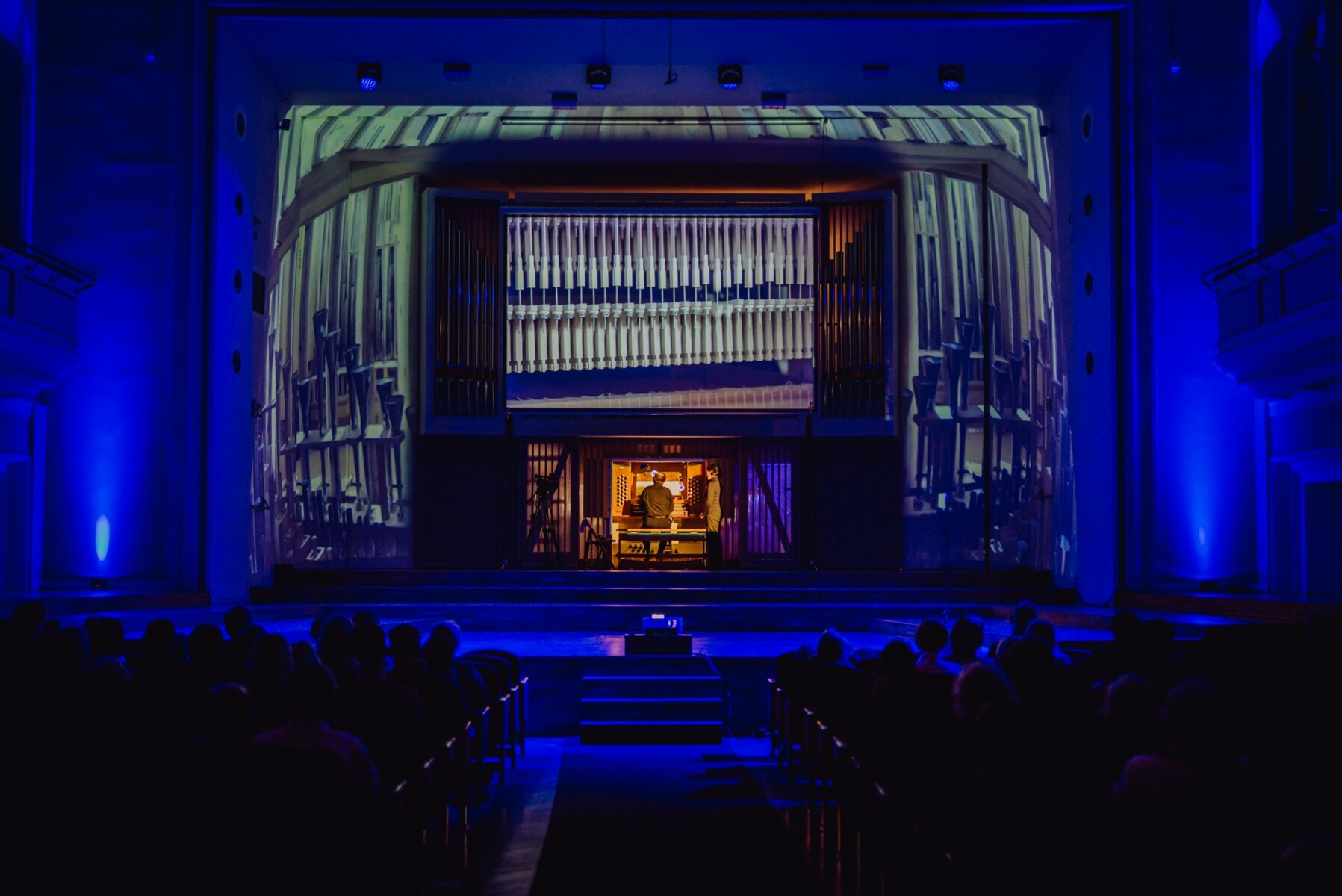 Zdjęcie przedstawia fragment widowni oraz scenę sali orkiestrowej. Na pierwszym planie znajduje się widownia filharmonii. Tyłem do widza, w fotelach siedzą słuchacze widowiska. Po środku znajduje się przejście pomiędzy siedzeniami, a na jego końcu przedstawiono schody które prowadzą do sceny. Zdjęcie jest ciemne, oświetlona jest scena filharmonii. Na scenie widoczna jest dwójka ludzi - osoba grająca na instrumencie muzycznym oraz stojąca obok, ustawiona bokiem do widowni. Nad muzykami wyświetla się obraz z rzutnika - fotografie instrumentów muzycznych. 