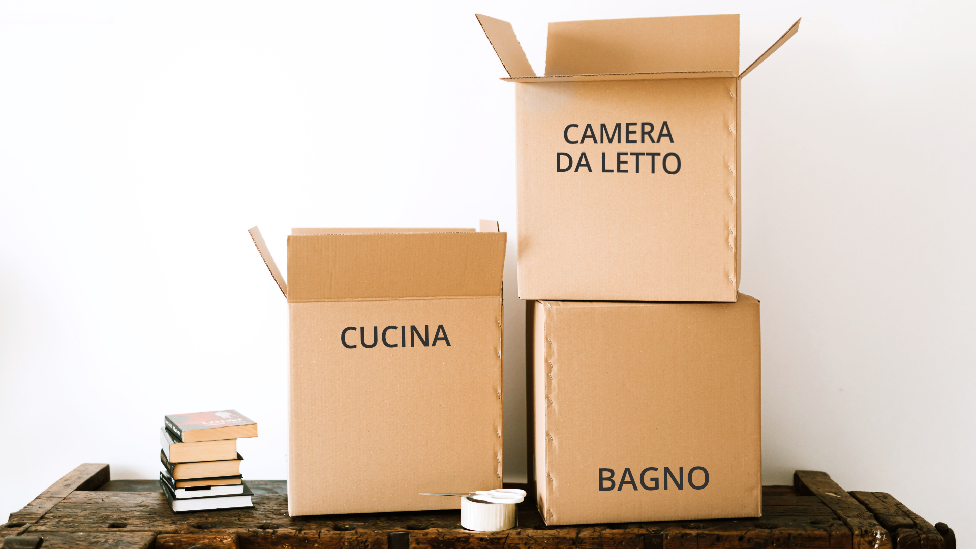 Zdjęcie przedstawia trzy kartonowe pudełka, które stoją na drewnianym blacie. Obok pudełek leży kilka książek. Na każdym pudle znajduje się napis. Odpowiednio: cucina, camera da letto, bagno.