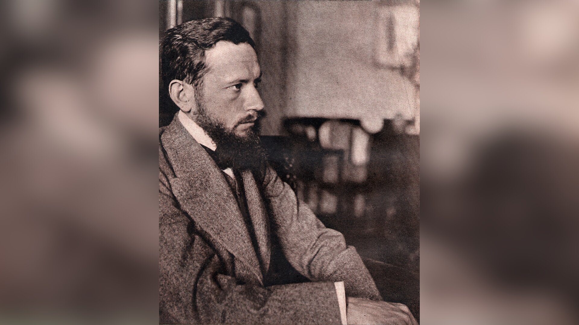 Zdjęcie przedstawia prawy profil siedzącego mężczyzny. To człowiek w sile wieku. Ma gładko zaczesane, krótkie włosy i gęstą brodę. Ubrany jest w kamizelkę i marynarkę.