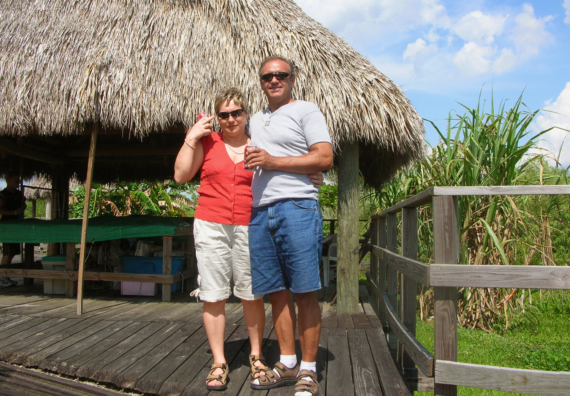 Fotografia przedstawia parę ludzi w wieku około 50 lat. Stoją na drewnianym tarasie, za nimi trzcinowe zadaszenie. Są objęci i uśmiechnięci.