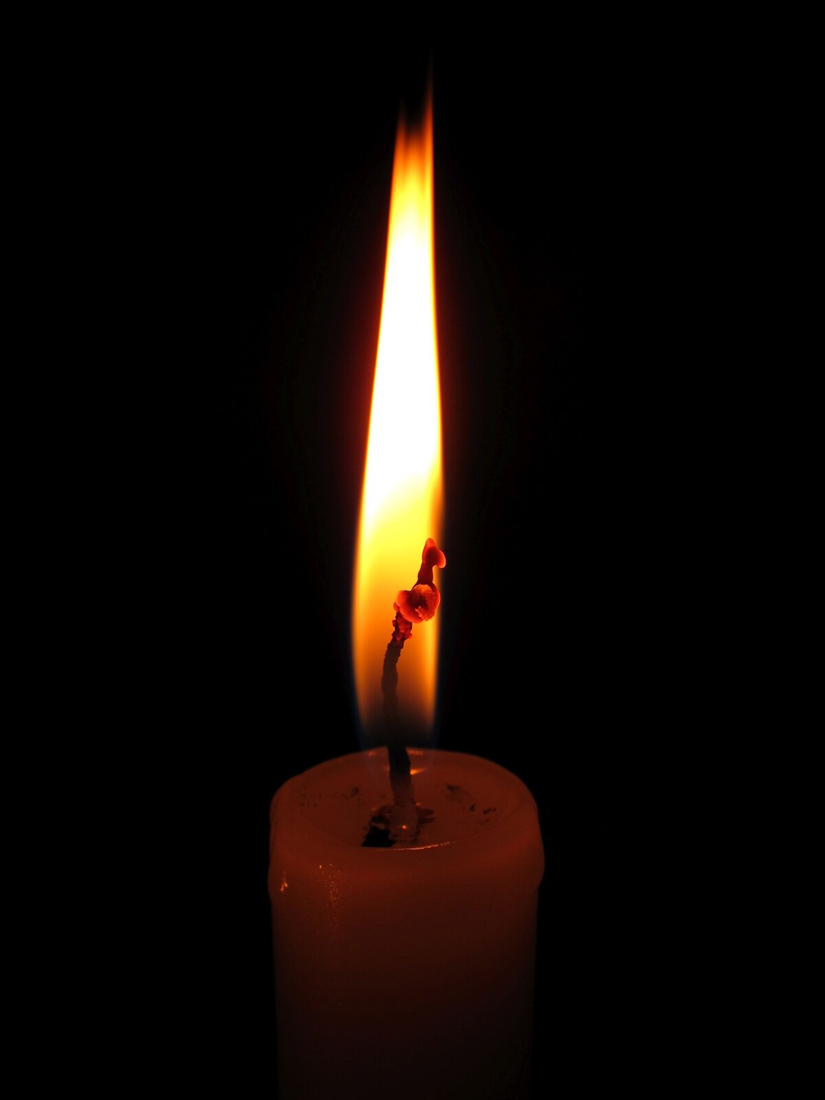 Ilustracja przedstawia palącą się świecę na czarnym tle. Jasny, długi płomień rozjaśnia świecę w górnej części. Na tle płomienia żarzy się knot.