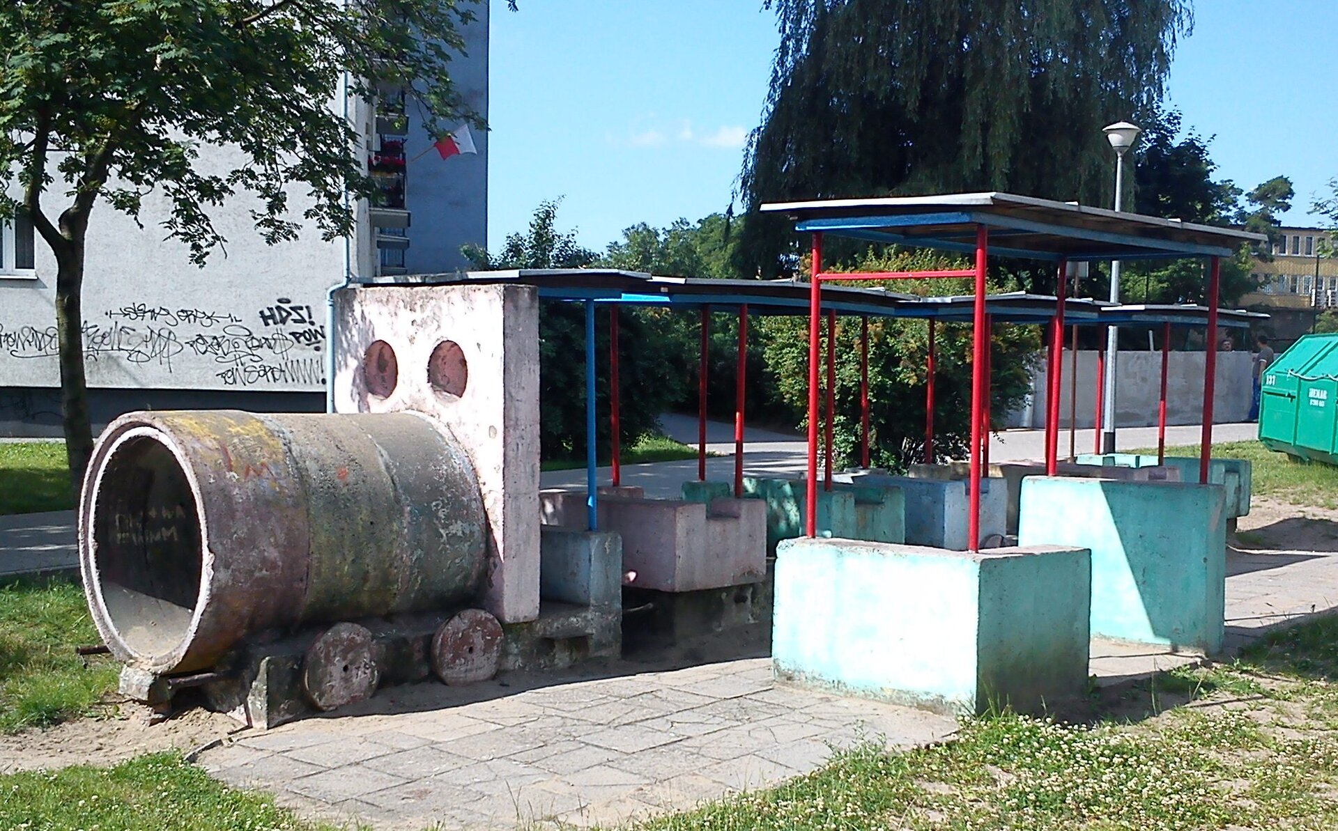 Fotografia przedstawia konstrukcję na placu zabaw na poznańskim osiedlu - pociąg stojący na stacji. Przedstawiony jest fragment przypominający szary przód lokomotywy – otwarta bryła walca na betonowej podstawie z elementami przypominającymi koła oraz jasnoróżowa ściana z dwoma otworami przypominającymi oczy. Następnie znajduje się kilka niskich murków w kolorze jasnoróżowym oraz turkusowym, z wychodzącymi od nich czerwonymi oraz niebieskimi belkami, łączące je z zadaszeniem. Podłoże przypomina jasne, kamienne płyty. Obok placu zabaw po lewej stronie widoczny jest trawnik, a za nim – drzewa o zielonych liściach, szary blok, niebieskie niebo, a także zielony kontener po prawej stronie.