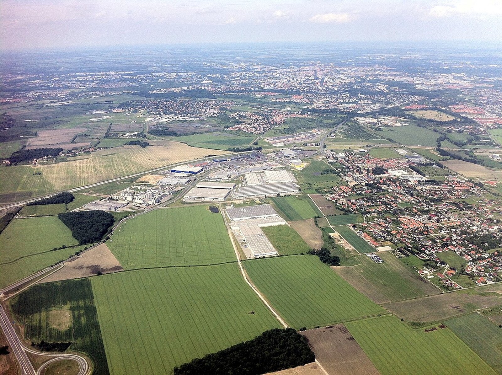 Lotnicze zdjęcie hal centrów logistycznych znajdujących się pod miastem. Hale mają po co najmniej kilkadziesiąt metrów długości, zbudowane na planie prostokąta, posiadają płaskie dachy. Są rozlokowane blisko siebie, jest ich kilka. Przed halami, w dolnej części ilustracji znajdują się pola uprawne, natomiast w górnej części, teren miejski lub zabudowa podmiejska.