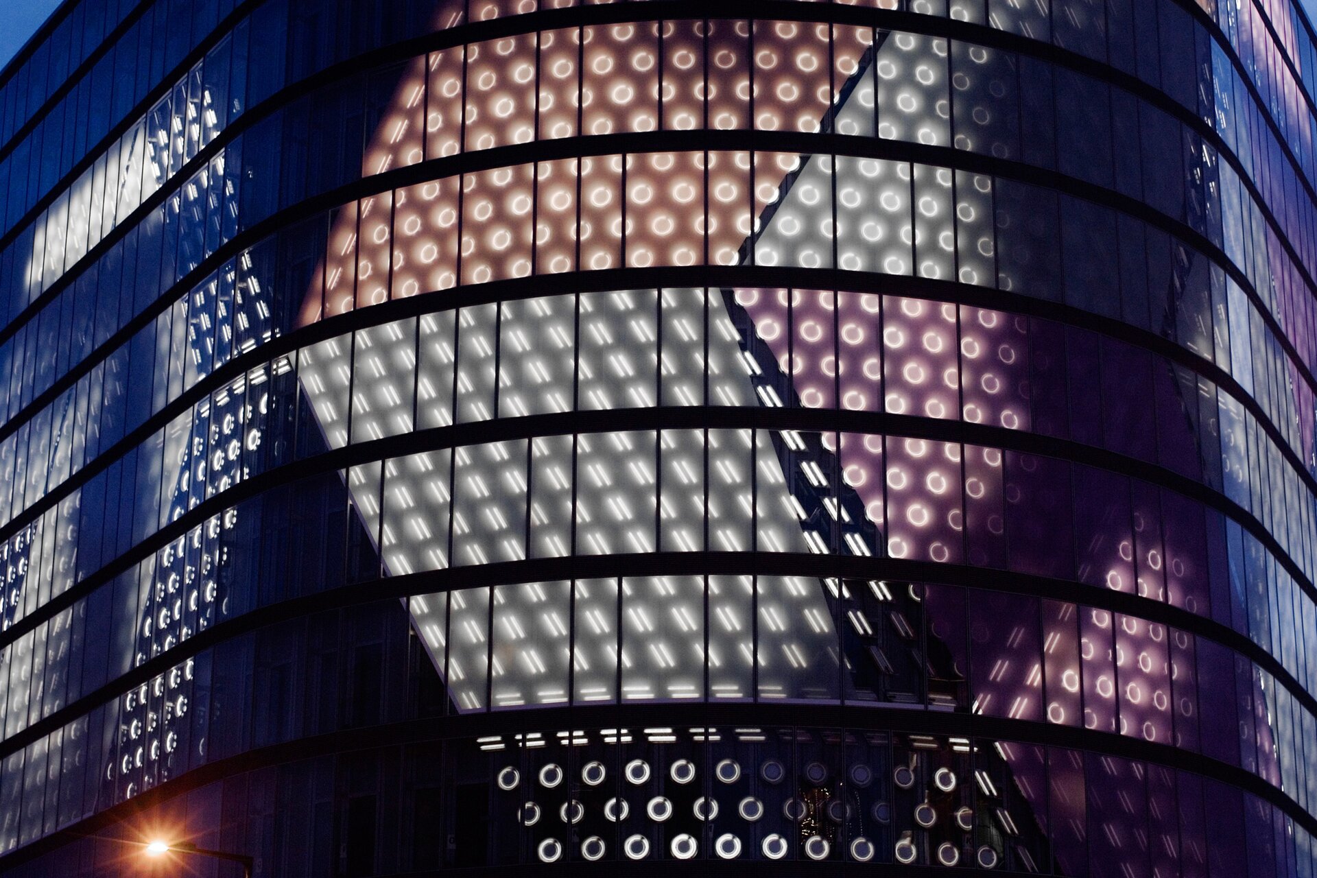 Fotografia przedstawia zbliżenie na podświetlony budynek. Ściana budynku ma kształt półokrągły, składa się z oddzielonych od siebie rzędów prostokątów przypominających szkło. Są one podświetlone różnymi kolorami, tworząc symetryczne, geometryczne wzory – zielone z cienkimi liniami ukośnymi, pomarańczowe z jasnymi okręgami, a także różowe i niebieskie. Na budynku widoczny jest także cień, jak również oświetlenie lampy z lewej strony.