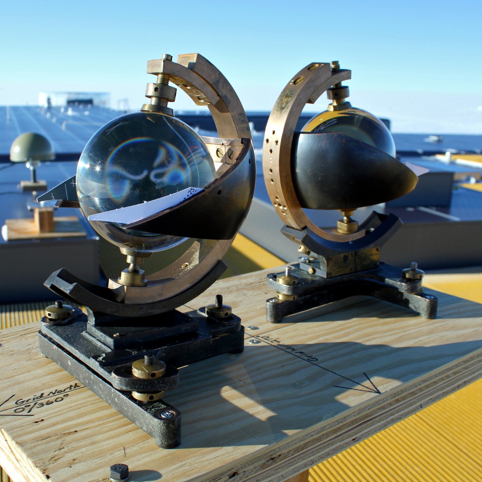 Zdjęcie przedstawia heliograf w kształcie szklanej kuli. Kula zawieszona jest na metalowym ramieniu w kształcie półksiężyca. Szkło kuli to soczewka.