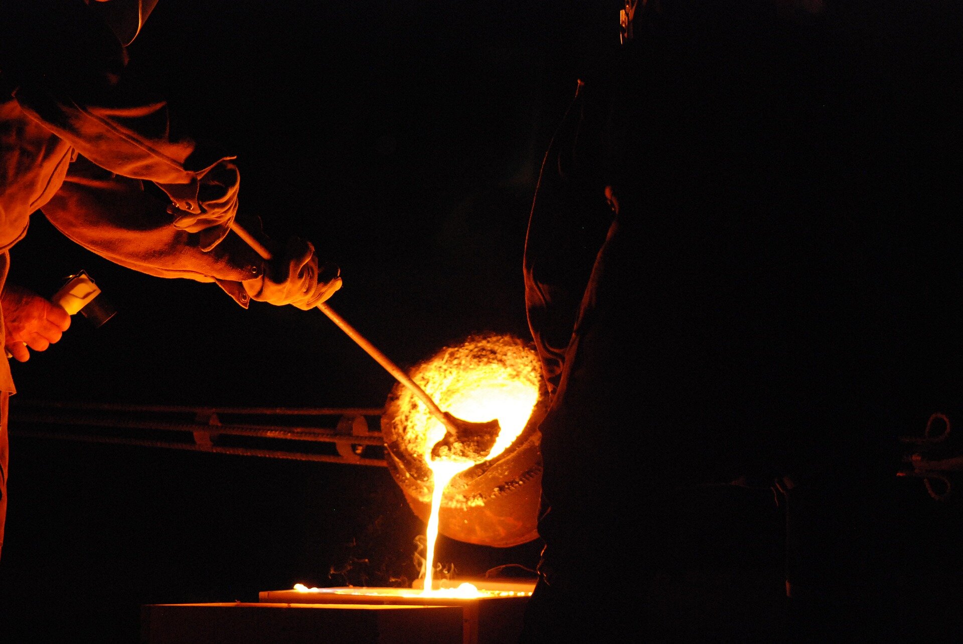 Zdjęcie przedstawia hutników pracujących przy wytopie stali. Z dużego tygla wylewana jest ciekła stal.