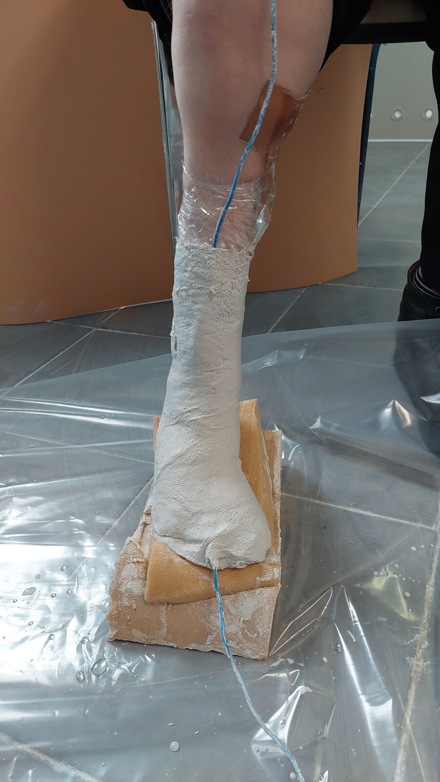 Zdjęcie przedstawia stopę owiniętą opaskami gipsowymi, stojącą na podłożu piankowym.