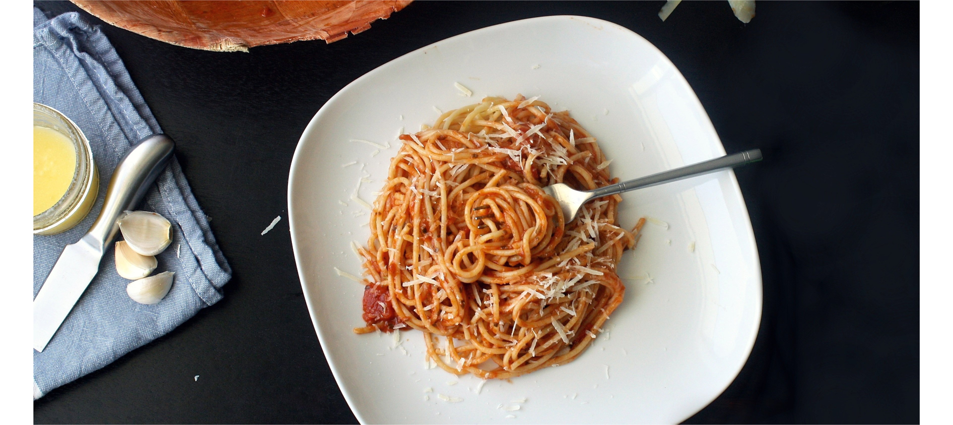 Zdjęcie przedstawia talerz, na którym znajduje się makaron z sosem pomidorowym. W talerzu leży widelec. Obok talerza widoczna jest serwetka na której leżą: ząbki czosnku i nóż. Zdjęcie zostało wykonane z góry.