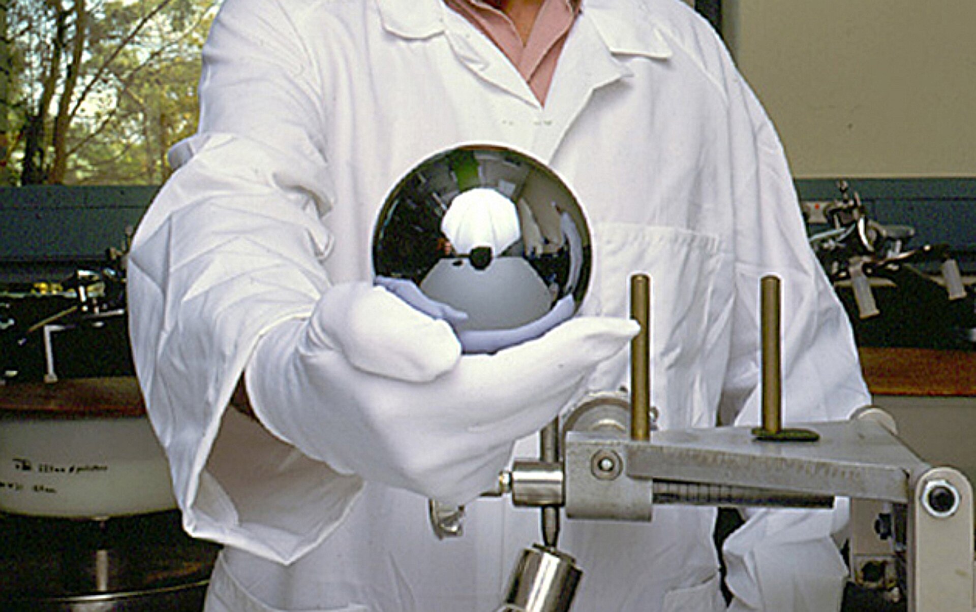 Zdjęcie przedstawia nowy wzorzec kilograma wykonany w ramach projektu Avogadro z monokryształu krzemu. Jest to jednocześnie najdoskonalsza wykonana przez człowieka kula na świecie, a jej średnica wynosi 93,75 mm. Idealnie gładka kula trzymana jest w dłoni w rękawiczce lateksowej przez naukowca, na tle pomieszczenia wypełnionego sprzętem badawczym.