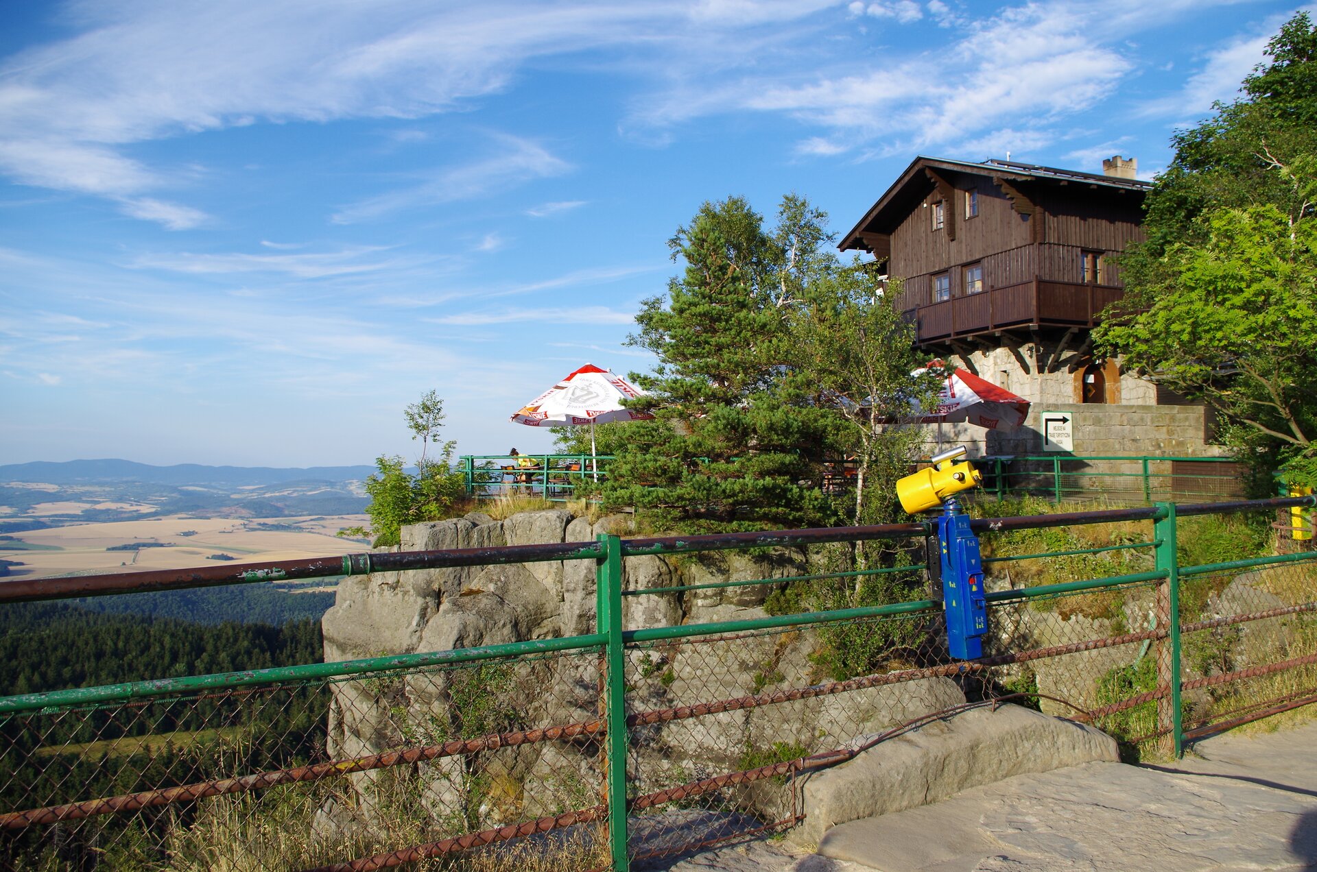 Na zdjęciu punkt widokowy zlokalizowany obok schroniska górskiego. Na pierwszym planie wysoka barierka, zamontowano na niej lunetę. Lewą część fotografii wypełnia obraz rozległego terenu znajdującego się u podnóża góry, pokrytego lasami lub polami uprawnymi. W tle linia masywu górskiego. Z prawej strony znajdujący się za barierką brązowy budynek schroniska, umieszczony jest na szarych skałach. Przed budynkiem ławki, krzesełka, parasole przeznaczone dla odwiedzających.