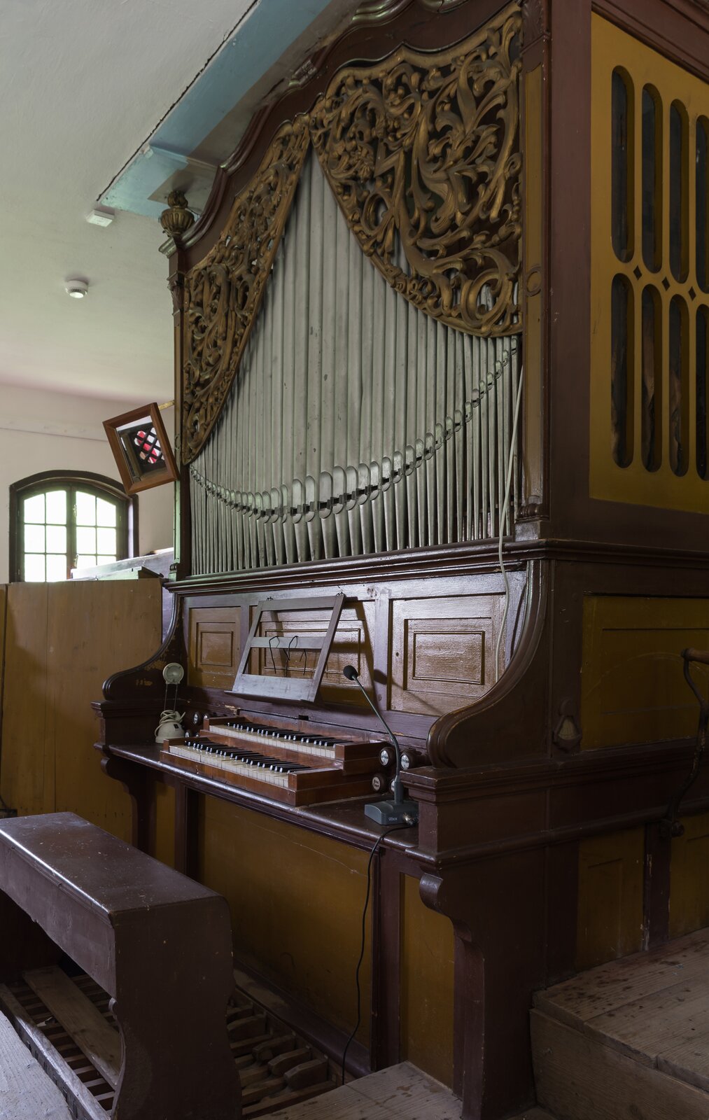 Fotografia przedstawia organy w kościele. Instrument jest drewniany, w górnej części ma złote zdobienia. Klawiatura ma czarne i białe klawisze. Nad częścią z klawiszami znajdują się długie metalowe rurki z otworami, ustawione obok siebie. Przed instrumentem stoi niewielka ława, pod którą znajdują się drewniane pedały. Obok klawiatury stoi mikrofon i lampka. W tle znajduje się okno.