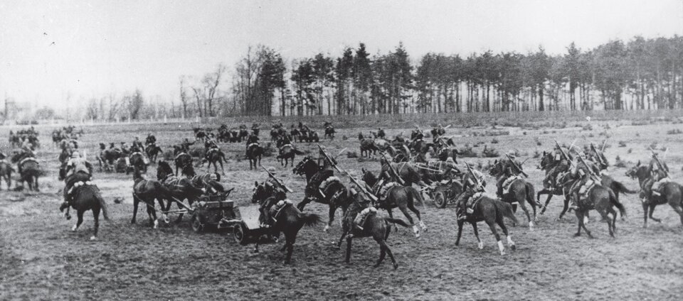 Zdjęcie biało‑czarne przedstawiające pluton artylerii przeciwpancernej 17 Pułku Ułanów Wielkopolskich podczas bitwy nad Bzurą we wrześniu 1939 r. Ułani jadą na czarnych koniach. Na drugim planie znajduje się niewielki las.
