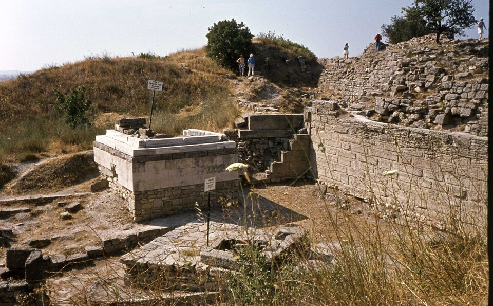 Zdjęcie przedstawia fragment wzgórza, na którym są ruiny antycznych budowli wykonanych z kamiennych bloków. W prawym rogu znajduje się mur obronny ułożony z niewielkich bloków kamiennych i kamieni. Wzdłuż muru przechadzają się turyści. W tle niewielkie krzaki i błękitne niebo.