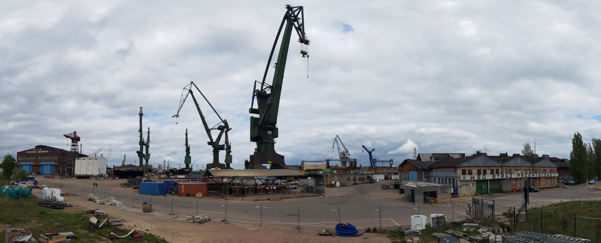 Zdjęcie przedstawia panoramę stoczni od strony lądu. Żurawie, budynki fabryczne, kontenery.