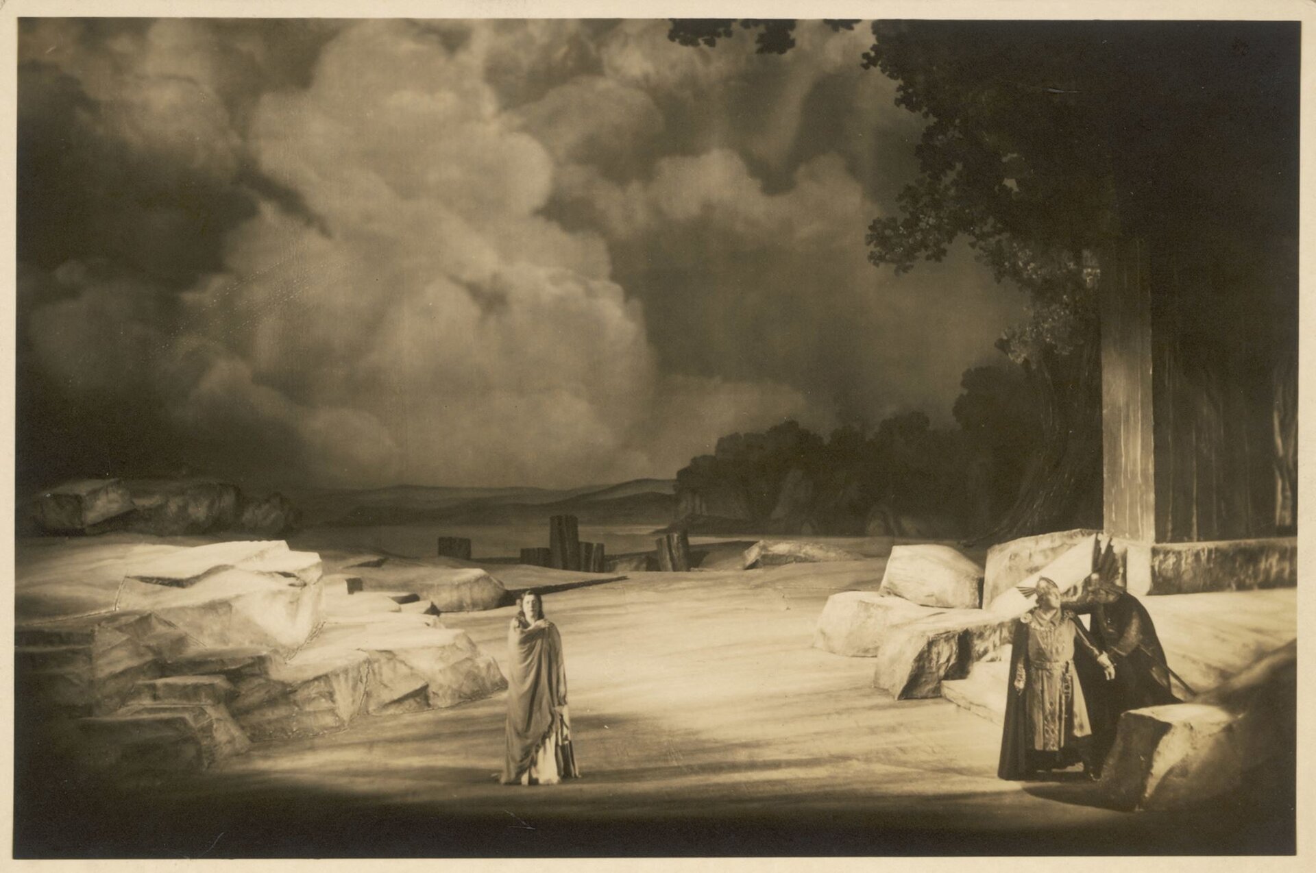 Ilustracja przedstawia realizację dramatu „Zmierzch bogów”, Bayreuth z 1938 r. Na zdjęciu znajduje się dwóch wojowników po prawej stronie, ubrani są w zbroje. W centrum stoi kobieta w jasnych szatach. W oddali widać skały, las i gęste, burzowe chmury.