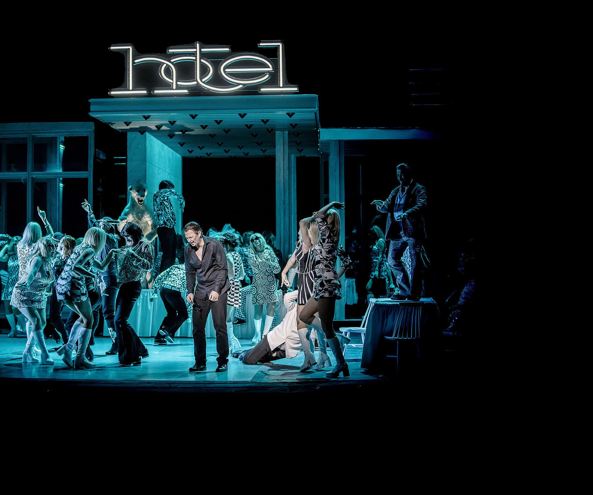 Zdjęcie przedstawia scenę z opery, ze współczesną scenografią. Scena przypomina dyskotekę, ludzie ubrani we wzorzyste stroje tańczą w różnych grupach. Niektórzy stoją na stołach. Na środku stoi zgarbiony mężczyzna, na którego pada białe światło. Reszta sceny skąpana jest w niebieskim, neonowym świetle. Za ludźmi znajduje się fragment budynku, na dachu którego znajduje się neonowy napis hotel.