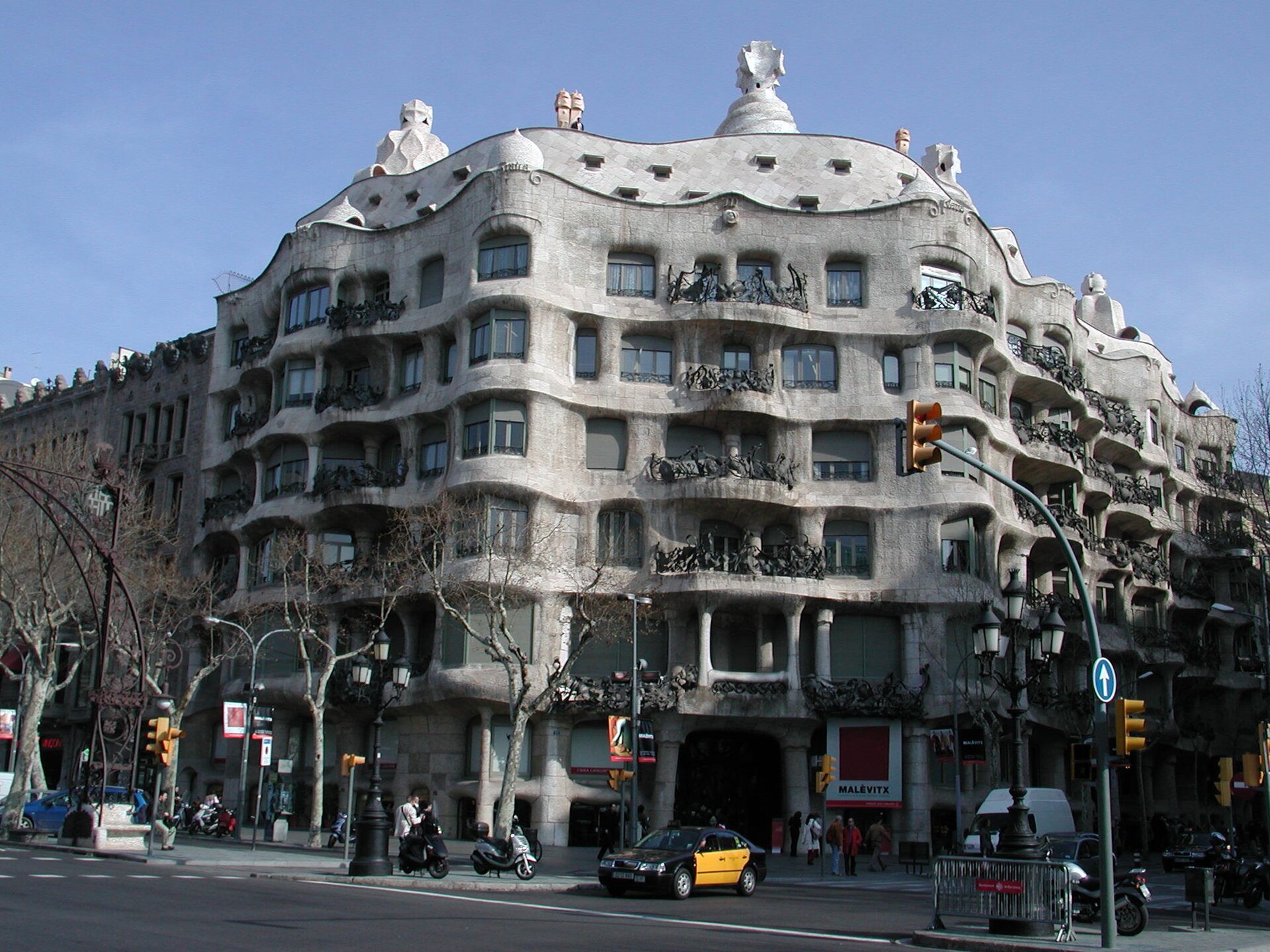 Ilustracja przedstawia budynek Casa Mila w Barcelonie zaprojektowany przez Antonia Gaudiego. Budynek jest wielokondygnacyjny i ma falisty kształt. Balkony zdobią kute z żelaza balustrady przybierające kształt wodorostów. Ozdobą fasady są również ptaki, które sprawiają wrażenie szykujących się do odlotu. Dach przypomina skorupę żółwia. Budynek znajduje się na skrzyżowaniu ulic.