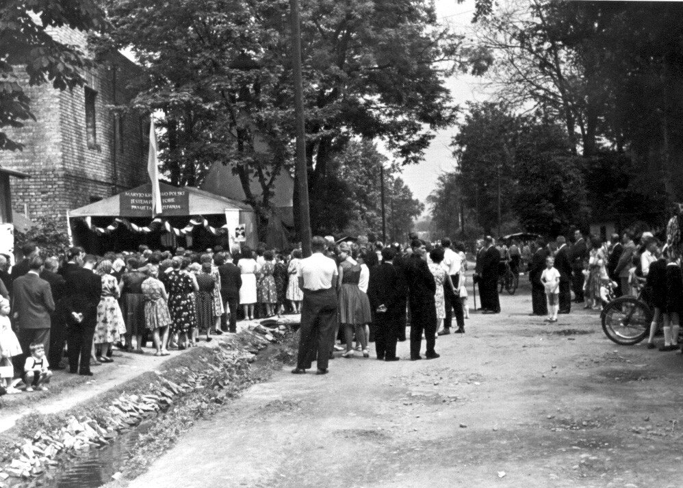 Kliknij, aby powiększyć. Zdjęcie przedstawia ulicę, na której stoją ludzie. Mają na sobie letnie ubrania - mężczyźni mają koszule z krótkimi rękawkami, kobiety noszą spódniczki i sukienki. Przy ścianie jednego z budynków po lewej stronie stoi kapliczka. Nad wejściem do kapliczki wiszą szarfy, na trójkątnym dachu widać napis. Przed kapliczką na chodniku stoi tłum ludzi. Wszyscy zwróceni w kierunku kapliczki. Na jezdni również znajdują się ludzie. Kilku z nich trzyma rowery. Widać dzieci w podkolanówkach i krótkich spodenkach. Wzdłuż ulicy stoją drzewa.