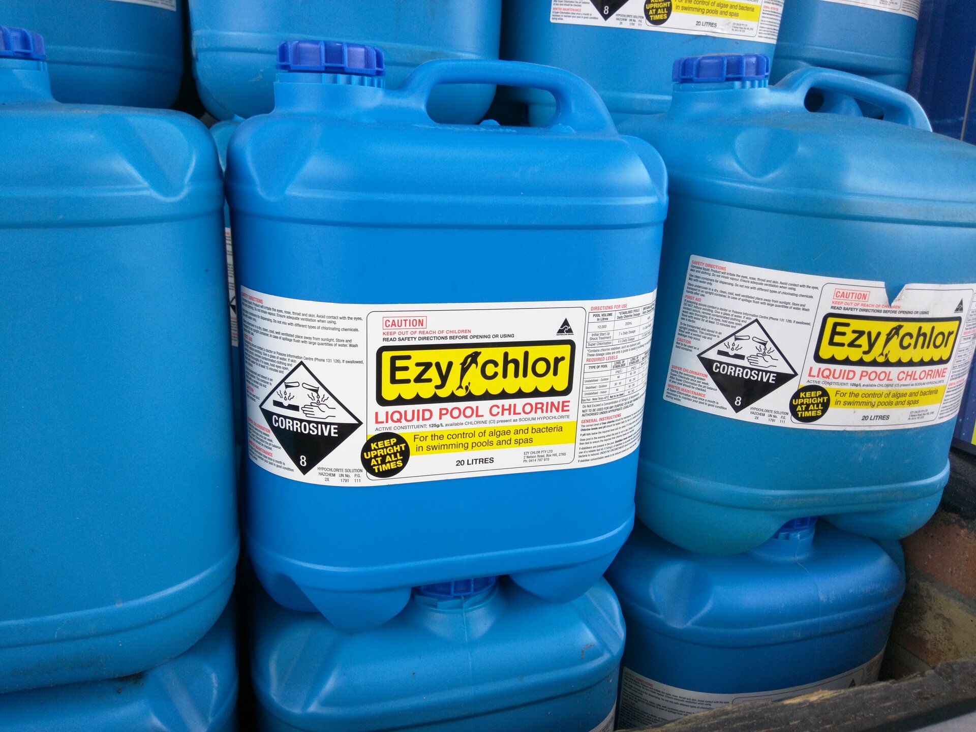 Zdjęcie przedstawia niebieskie kanistry wypełnione płynem do odkażania wody. Na etykiecie znajduje się zapis: liquid pool chlorine.