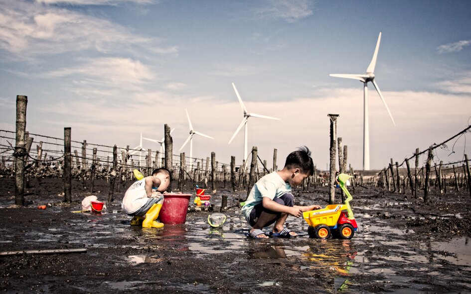 Zdjęcie przedstawia degradację środowiska naturalnego w Chinach, która znacznie ograniczyła dostęp do wody pitnej. Dzieci bawią się w błocie na opustoszałym polu. W pobliżu wiatraki, kumulujące energię z wiatru.