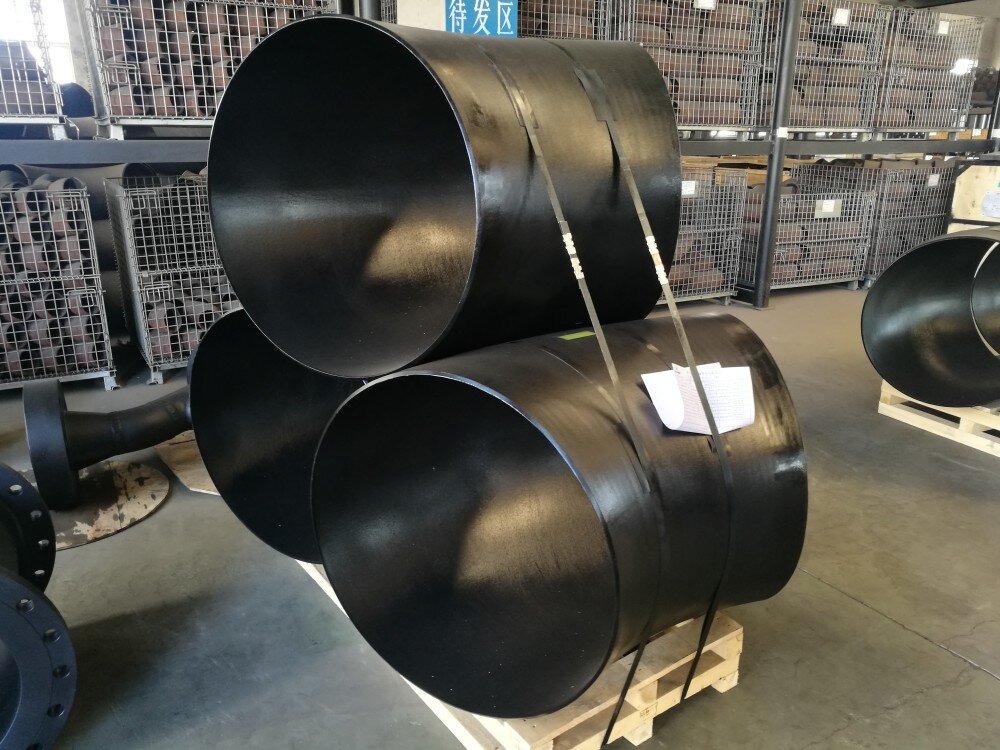 Zdjęcie przedstawia przygotowanie kolan PVC do transportu. Widać trzy duże kolana, które mają postać lekko zgiętych rur. Są one ułożone na palecie, dwa stanowią podstawę, trzecie znajduje się na górze. Kolana są przymocowane do palety pasami.