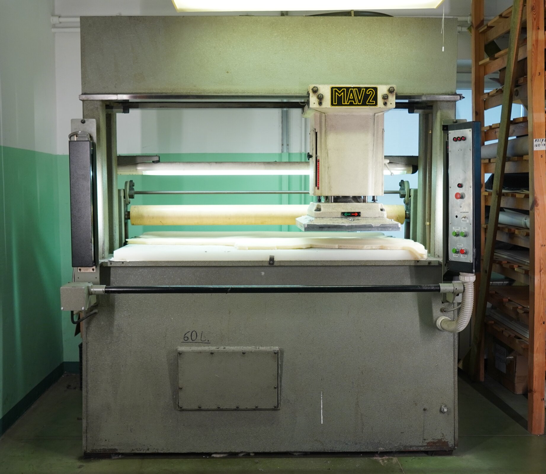 Zdjęcie przedstawia dużą szarą wolnostojącą maszynę. W jej tyle znajduje się szeroka prasa, a z przodu wąska czarna rurka. Pośrodku, po prawej stronie, znajduje się zawieszony element. Nagranie tożsame z treścią.