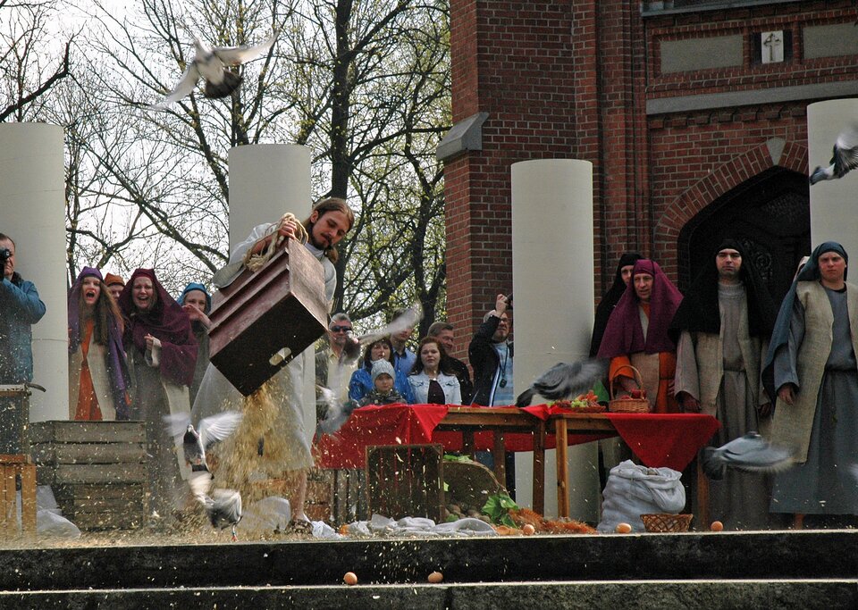 Kolorowe zdjęcie ukazuje fragment przedstawienia odbywającego się przed kościołem, na tle drzew i ściany z czerwonej cegły. Na pierwszym planie znajduje się mężczyzna w białej szacie, w średnim wieku, który wysypuje zboże ze skrzynki. Na ziemi leżą porozrzucane warzywa, kosze. W powietrzu latają gołębie. Mężczyźnie przygląda się grupa aktorów w długich szatach, w chustach na głowach, którzy wyrażają niezadowolenie, że mężczyzna niszczy ich produkty, które wystawili na sprzedaż.