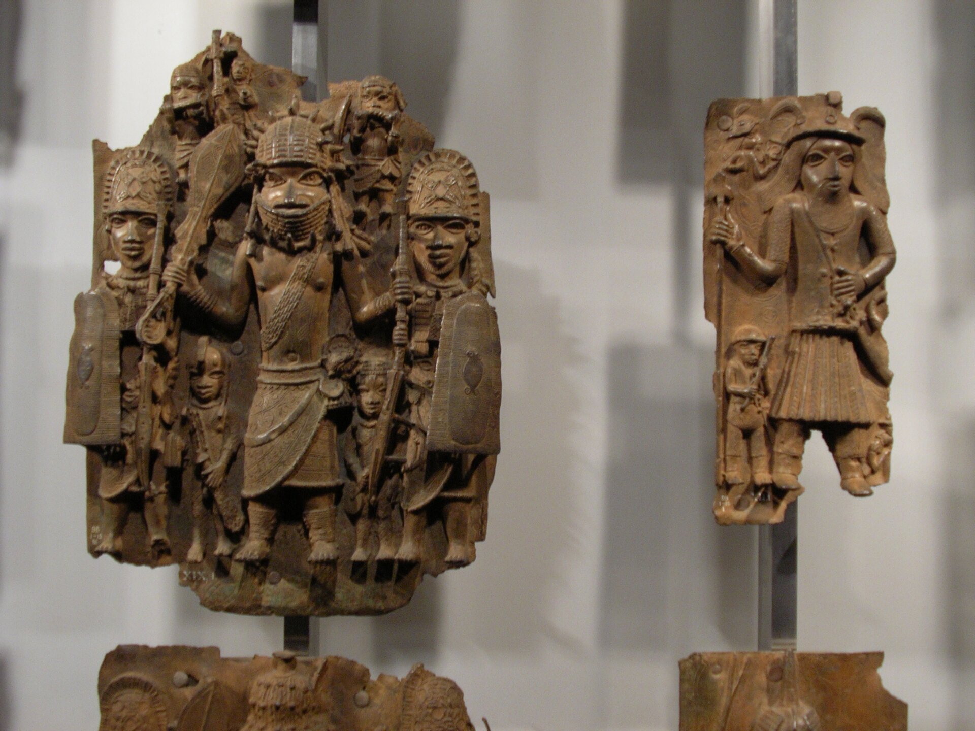 Zdjęcie z muzeum przedstawia dwie plakiety z brązu. Pierwsza z lewej przedstawia w części środkowej wojownika przepasanego tuniką i pasem przez klatkę piersiową od lewego barku do prawego biodra. W prawej dłoni dzierży obiekt o formie maczugi, w lewej zaś wąski, podłużny miecz w pochwie. Po obu jego stronach dwóch wojowników z afrykańskimi hełmami i prostokątnymi tarczami. W głębi niżsi wojownicy, sięgający pierwszym postaciom wysokością do bioder. Po prawej stronie druga plakieta, przedstawiająca mężczyznę z brodą, płaskim nakryciem głowy, laską w prawej dłoni. Lewą trzyma na pasku, przewieszonym przez prawe ramię do lewego biodra. Plakieta nosi znamiona uszkodzeń, jest niepełna, nie widać nóg postaci.  