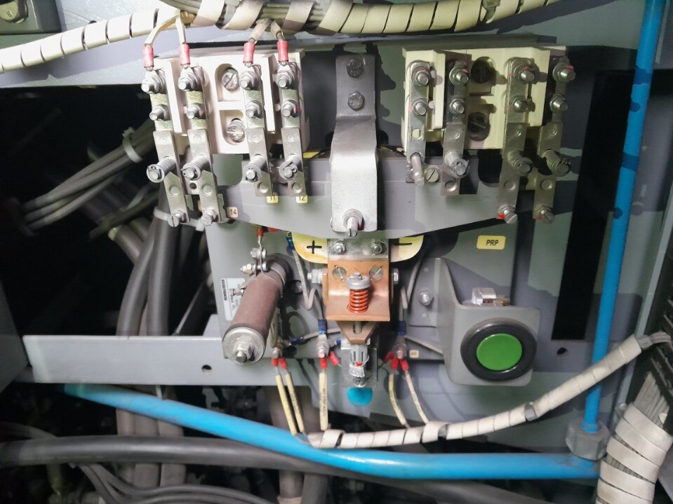 Zdjęcie przedstawia przekaźnik różnicowy. Na ścianie zamocowane jest urządzenie z wieloma wystającymi śrubami. Do niektórych przymocowane są przewody elektryczne. W dolnym prawym rogu znajduje się duży zielony przycisk. Po lewej stronie widoczny jest cylinder.