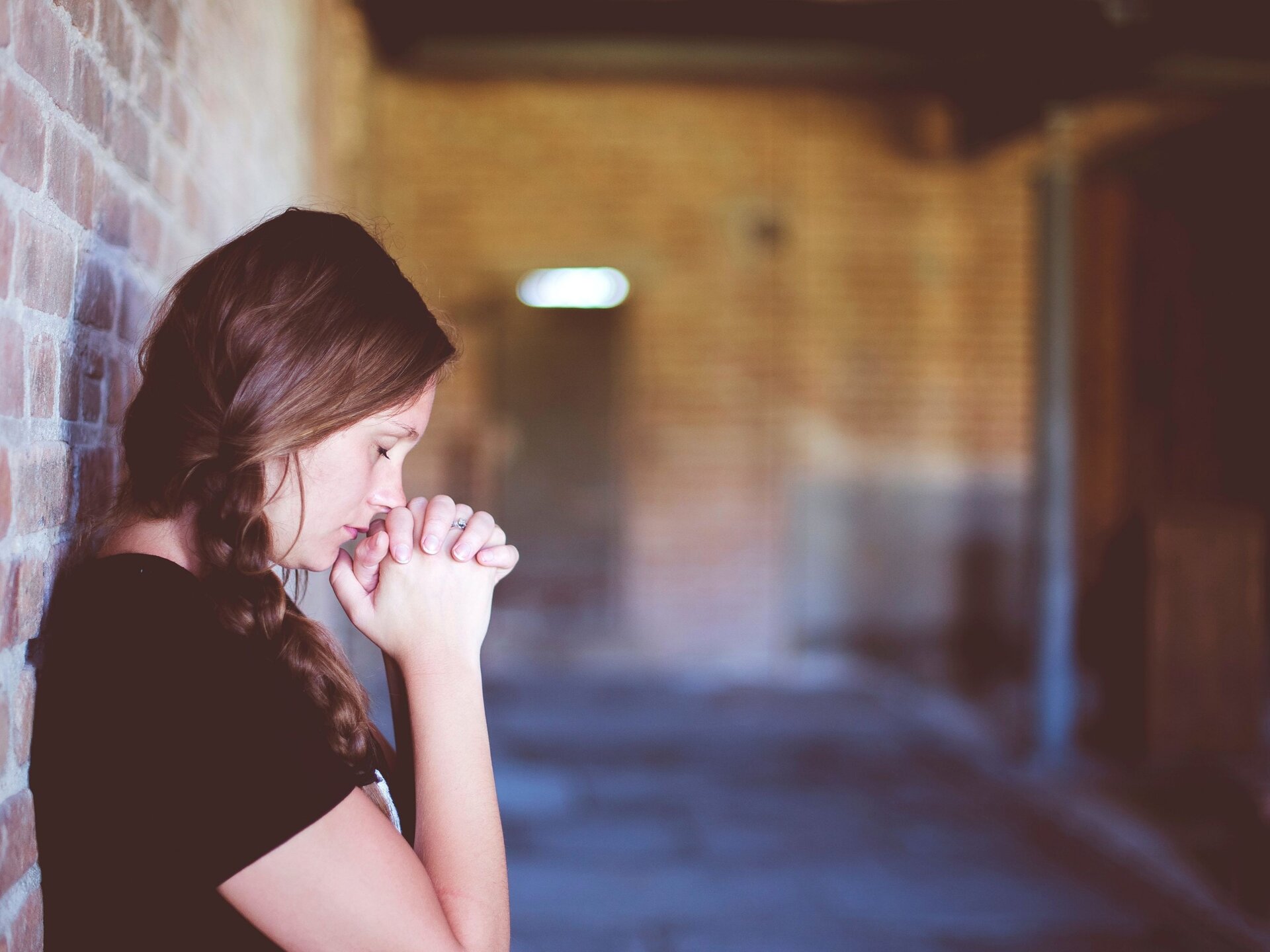 Ilustracja numer cztery. Zdjęcie przedstawia młodą dziewczynę z warkoczem opartą o ścianę z cegieł. Dziewczyna sfotografowana jest z prawego profilu. Ma splecione dłonie podniesione na wysokość ust i zamknięte oczy.  